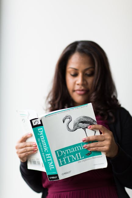 A woman with dark hair, dressed in a black jacket and maroon top, is holding a book titled 'Dynamic HTML' featuring a flamingo illustration on the cover. She is standing indoors against a plain white