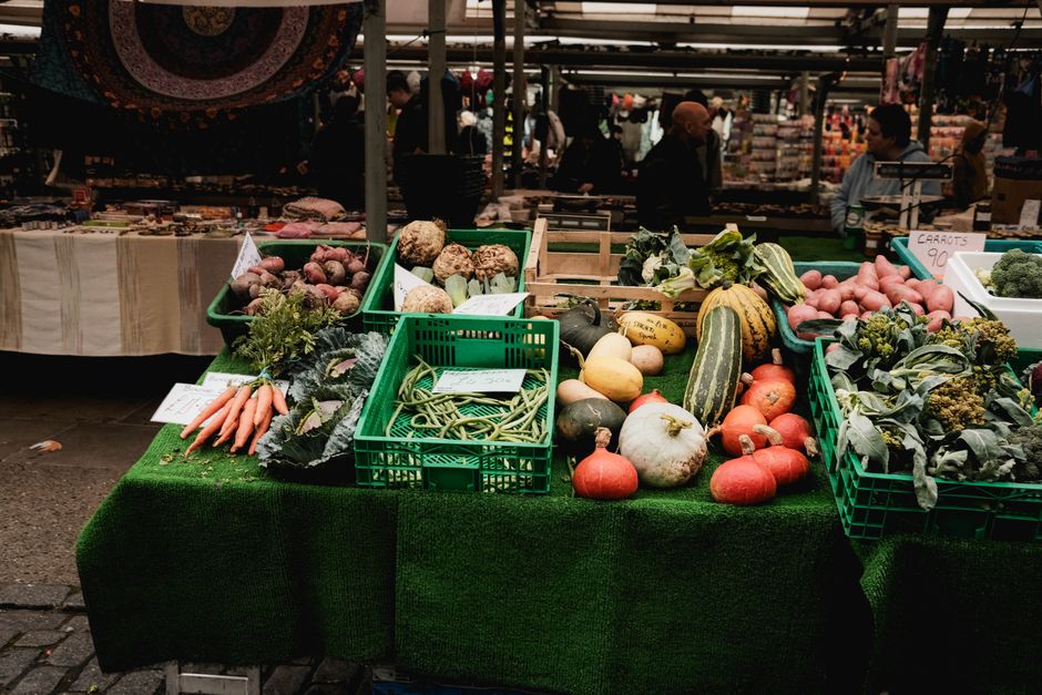 A market stall at Maltby Street Bermondsey market showing an assortment of fresh vegetables and produce on a green artificial grass tablecloth. Visible items include carrots, potatoes, squash, broccol